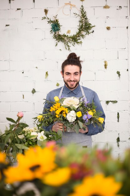 Lächelnder junger männlicher Florist, der in der Hand den Blumenblumenstrauß gegen Ziegelsteinweißwand hält