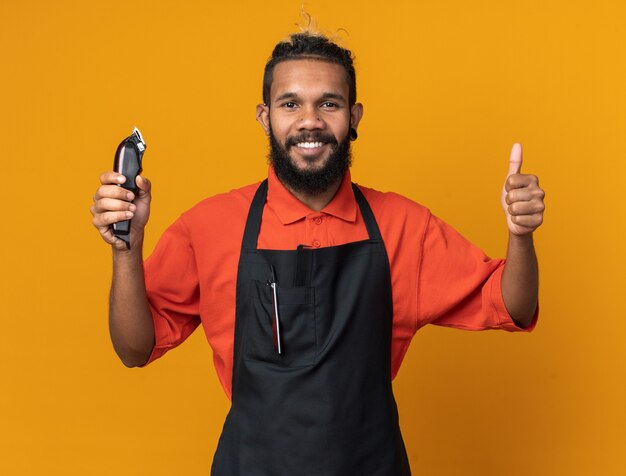 Lächelnder junger männlicher Friseur in Uniform, der Haarschneidemaschinen hält und nach vorne schaut, der Daumen nach oben isoliert auf oranger Wand zeigt