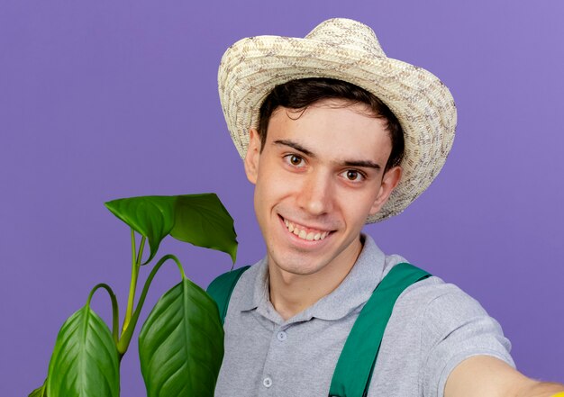 Lächelnder junger männlicher Gärtner, der Gartenhut trägt, steht mit Pflanze, die vorgibt, Kamera zu halten