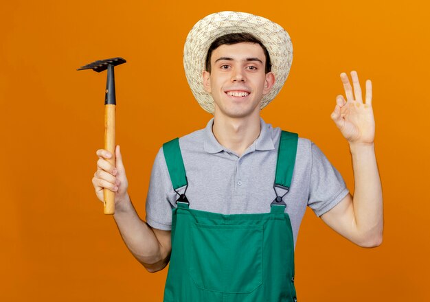 Lächelnder junger männlicher Gärtner mit Gartenhut gestikuliert ok Handzeichen und hält Rechen