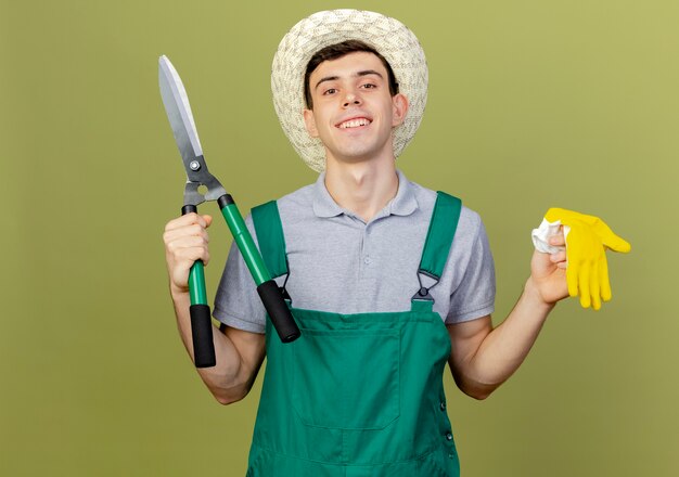 Lächelnder junger männlicher Gärtner mit Gartenhut hält Klipper und Handschuhe