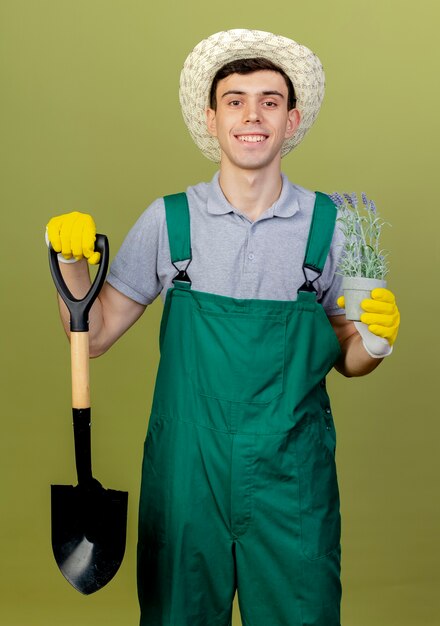 Lächelnder junger männlicher Gärtner mit Gartenhut hält Spaten und Blumen im Blumentopf