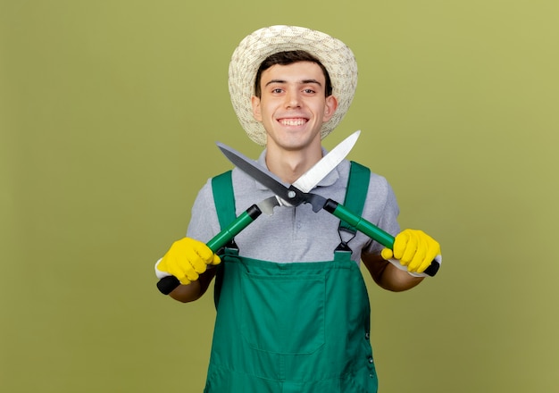 Lächelnder junger männlicher gärtner mit gartenhut und handschuhen hält schermaschinen