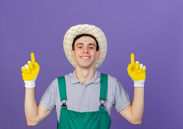 Lächelnder junger männlicher Gärtner mit Gartenhut und Handschuhen zeigt nach oben
