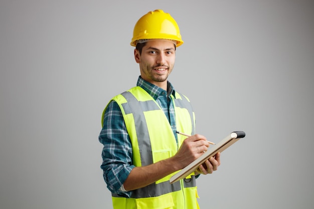 Lächelnder junger männlicher Ingenieur in Uniform, der in der Profilansicht mit Bleistift und Notizblock steht, während er die Kamera isoliert auf weißem Hintergrund mit Kopierraum betrachtet