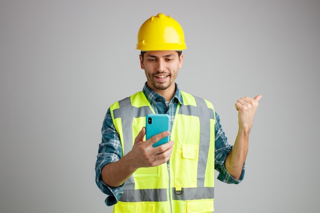 Lächelnder junger männlicher Ingenieur mit Schutzhelm und Uniform, der das Handy hält und betrachtet, das zur Seite zeigt, isoliert auf weißem Hintergrund