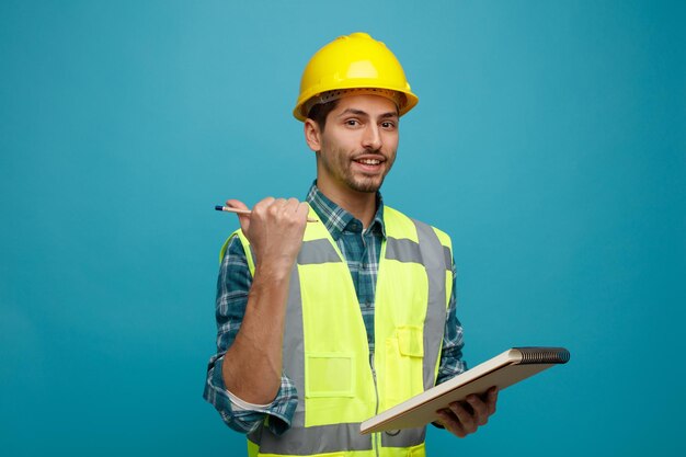 Lächelnder junger männlicher Ingenieur mit Schutzhelm und Uniform mit Bleistift und Notizblock, der auf die Kamera zeigt, die auf die Seite zeigt, die isoliert auf blauem Hintergrund steht