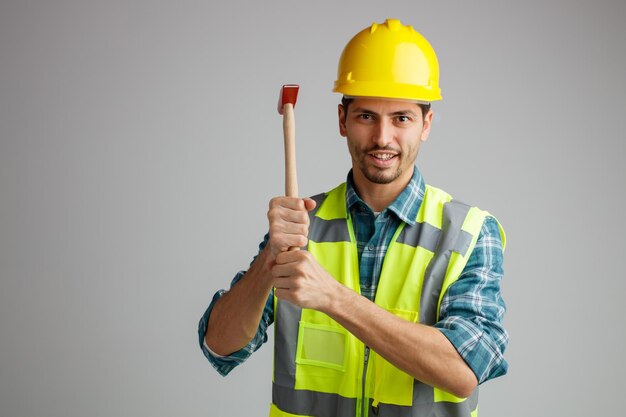 Lächelnder junger männlicher Ingenieur mit Schutzhelm und Uniform mit Hammer, der isoliert auf weißem Hintergrund mit Kopierraum in die Kamera blickt