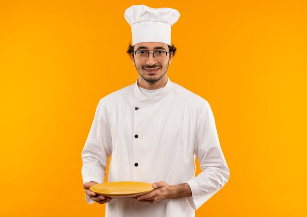 Lächelnder junger männlicher Koch, der die Kochuniform und die Brille hält, die Platte lokalisiert auf gelber Wand tragen