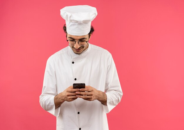 Lächelnder junger männlicher Koch, der Kochuniform und Brille wählt Nummer am Telefon lokalisiert auf rosa Wand