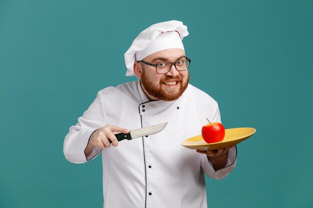 Lächelnder junger männlicher Koch mit einheitlicher Brille und Kappe, der einen Teller mit Apfel darin und Messer hält, der isoliert auf blauem Hintergrund in die Kamera blickt