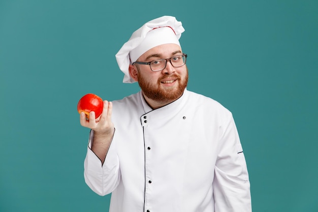 Lächelnder junger männlicher Koch mit einheitlicher Brille und Mütze mit Apfel, der isoliert auf blauem Hintergrund in die Kamera blickt