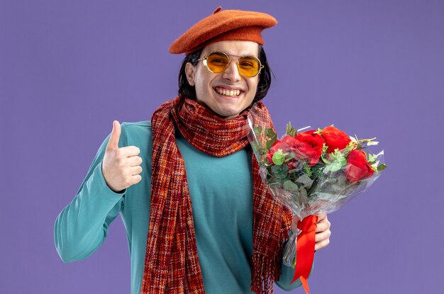 Lächelnder junger Mann am Valentinstag mit Hut mit Schal und Brille, der einen Blumenstrauß hält, der den Daumen einzeln auf blauem Hintergrund zeigt