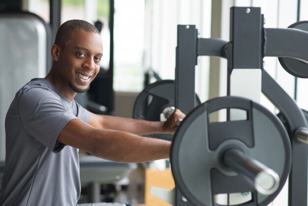 Lächelnder junger Mann, der am Barbell steht und Kamera betrachtet