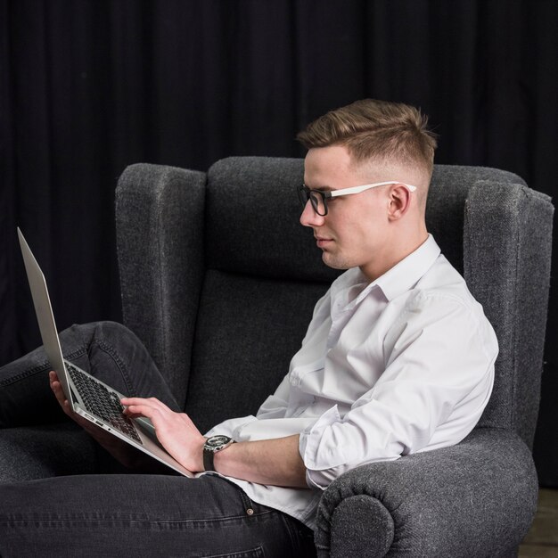 Lächelnder junger Mann, der auf dem Lehnsessel unter Verwendung des Laptops sitzt