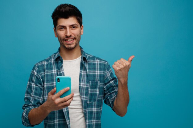 Lächelnder junger Mann, der ein Handy hält und in die Kamera blickt, die auf die Seite zeigt, die auf blauem Hintergrund isoliert ist