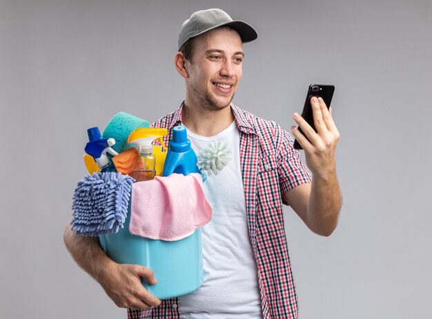 Lächelnder junger Mann, der eine Kappe trägt, die einen Eimer mit Reinigungswerkzeugen hält und das Telefon in seiner Hand isoliert auf weißer Wand betrachtet