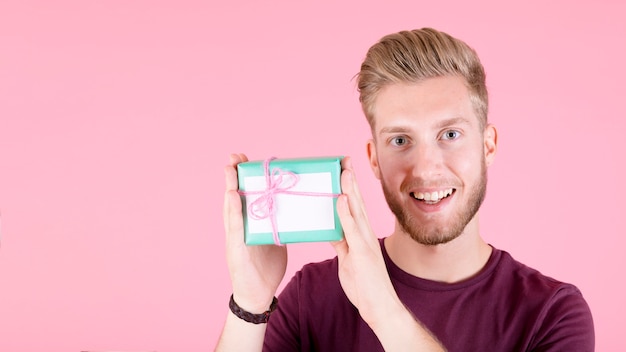 Lächelnder junger Mann, der Geschenkbox gegen rosa Hintergrund zeigt