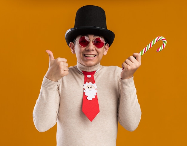 Kostenloses Foto lächelnder junger mann, der hut mit weihnachtskrawatte und brille trägt, die weihnachtsbonbon hält, der daumen oben auf gelber wand zeigt