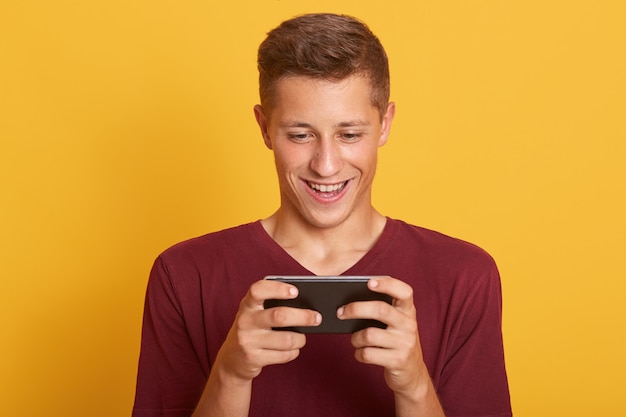 Kostenloses Foto lächelnder junger mann, der spiel auf smartphone spielt, sieht glücklich und konzentriert aus und schaut lächelnd auf den bildschirm seines geräts