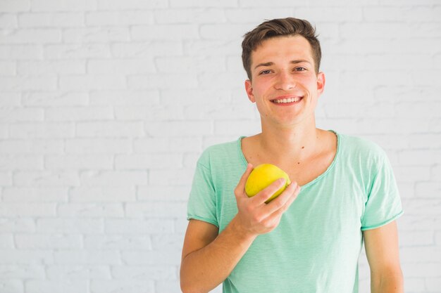 Lächelnder junger Mann, der vor der Wand hält grünen Apfel steht