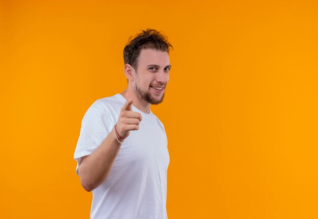 Lächelnder junger Mann, der weißes T-Shirt trägt, das Sie Geste auf lokalisiertem orange Hintergrund zeigt