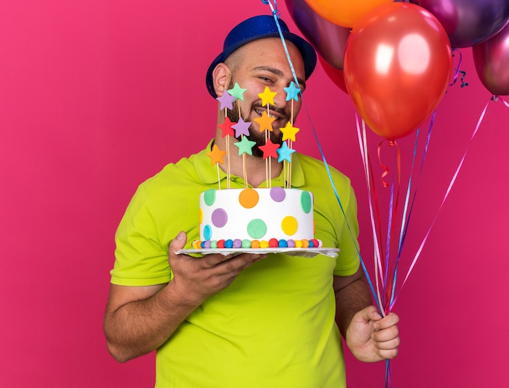 Kostenlose Foto lächelnder junger mann mit blauem partyhut, der luftballons mit kuchen hält