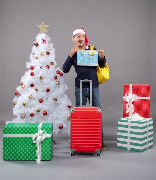 lächelnder junger Mann mit gelbem Rucksack, der Karte mit beiden Händen nahe Weihnachtsbaum auf grau hält