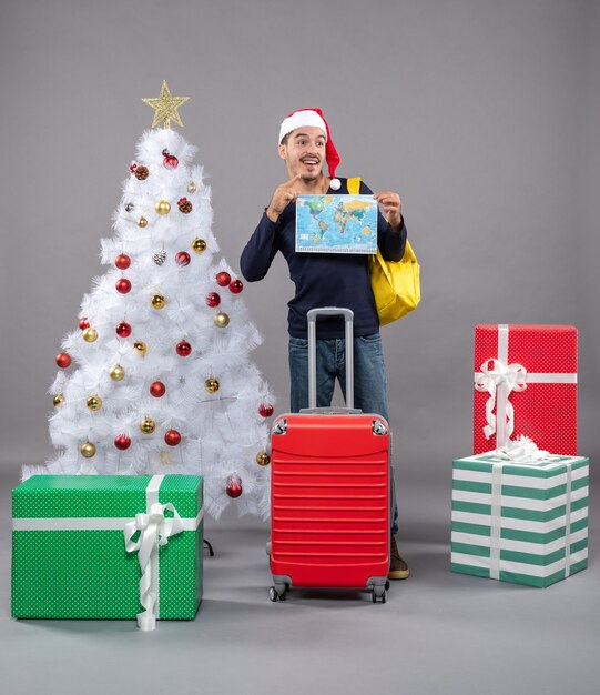 lächelnder junger Mann mit gelbem Rucksack, der Karte mit beiden Händen nahe Weihnachtsbaum auf grau hält