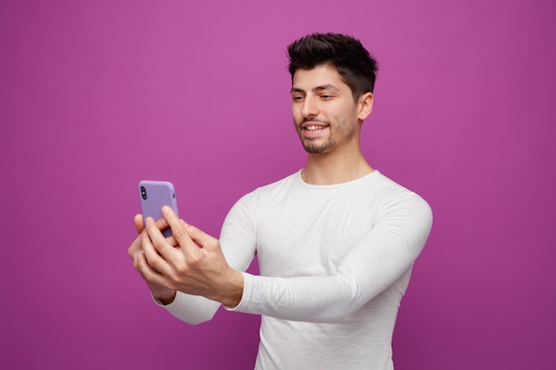 Lächelnder junger mann mit handy, der ein selfie isoliert auf violettem hintergrund macht