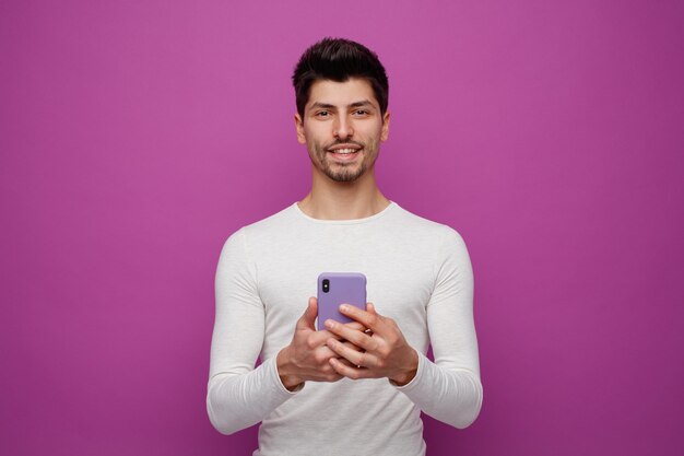 Lächelnder junger Mann mit Handy, der isoliert auf violettem Hintergrund in die Kamera blickt