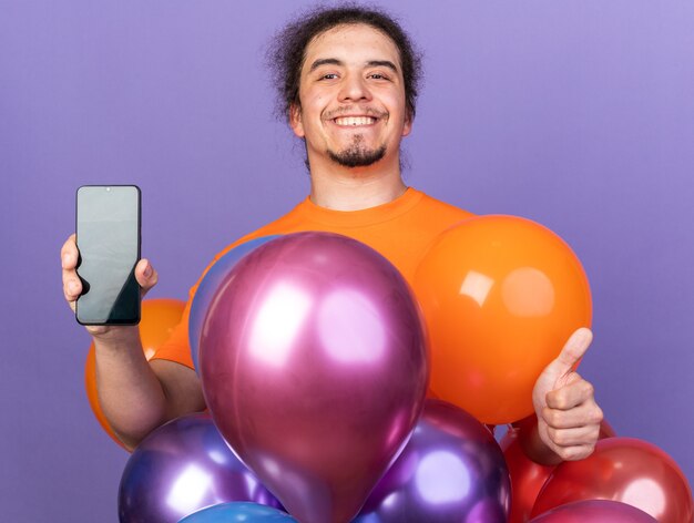 Lächelnder junger Mann mit orangefarbenem T-Shirt, der hinter Luftballons steht und das Telefon hält, das Daumen nach oben zeigt, isoliert auf lila Wand