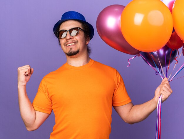 Lächelnder junger Mann mit Partyhut mit Brille, der Luftballons hält, die eine Ja-Geste einzeln auf lila Wand zeigen