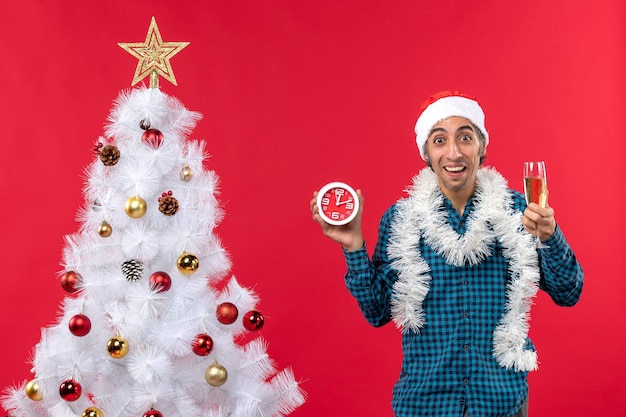 Lächelnder junger mann mit weihnachtsmannhut und halten eines glases wein und der uhr, die nahe weihnachtsbaum auf rot stehen