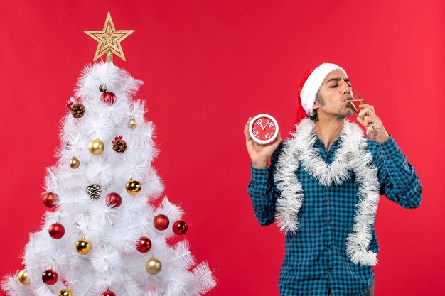 Lächelnder junger Mann mit Weihnachtsmannhut und Trinken eines Glases Wein und Halten der Uhr, die nahe Weihnachtsbaum auf Rot steht