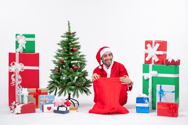 Lächelnder junger Mann verkleidet als Weihnachtsmann mit Geschenken und geschmücktem Weihnachtsbaum, der auf dem Boden sitzt, der für Kamera auf weißem Hintergrund aufwirft