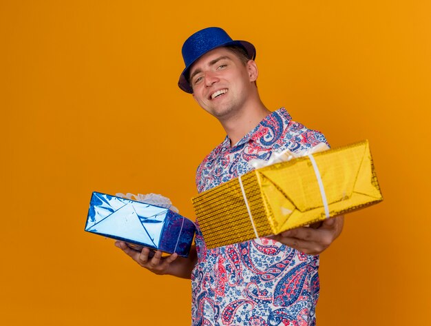 Lächelnder junger Party-Typ, der blauen Hut trägt, der Geschenkboxen lokalisiert auf Orange hält