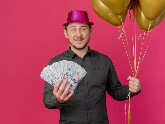 Lächelnder junger Party-Typ, der rosa Hut hält, der Luftballons mit Bargeld lokalisiert auf Rosa hält