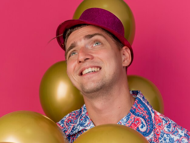 Lächelnder junger Party-Typ, der rosa Hut trägt, der unter Luftballons steht, die auf rosa isoliert werden