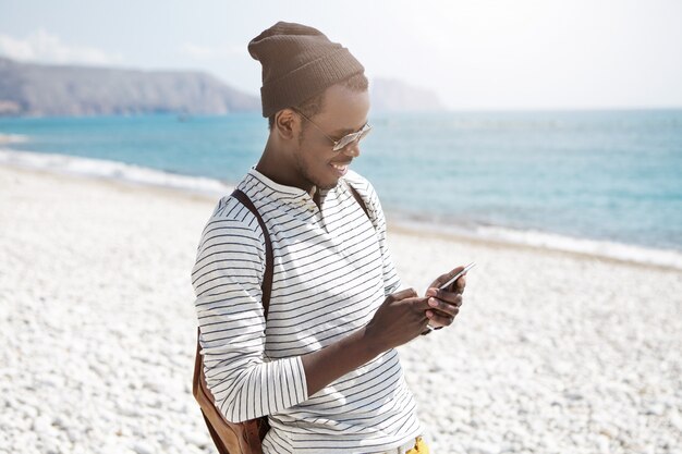 Lächelnder junger schwarzer europäischer männlicher Tourist in Hut und Schatten, der 3g Internet auf Handy am Strand benutzt, Bilder über soziale Medien teilt, glückliche Tage während seiner Sommerferien am Meer genießt