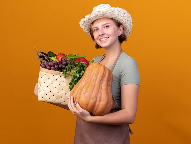 Lächelnder junger slawischer weiblicher Gärtner, der Gartenhut hält, der Gemüsekorb und Kürbis auf Orange hält