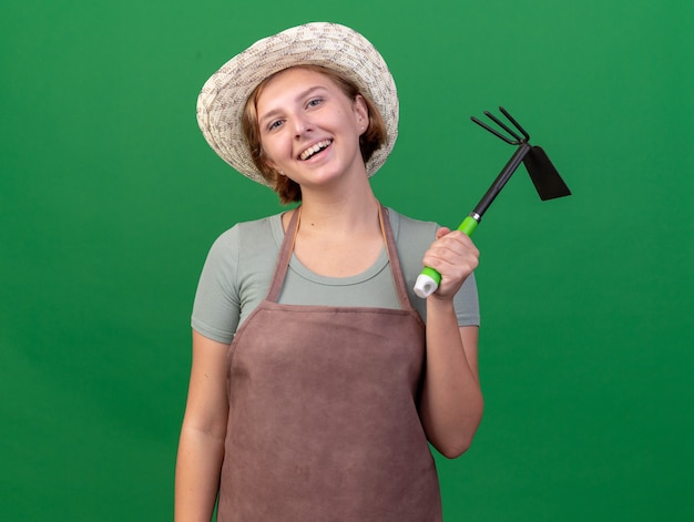 Lächelnder junger slawischer weiblicher Gärtner, der Gartenhut hält, der Hacke Rechen auf Grün hält