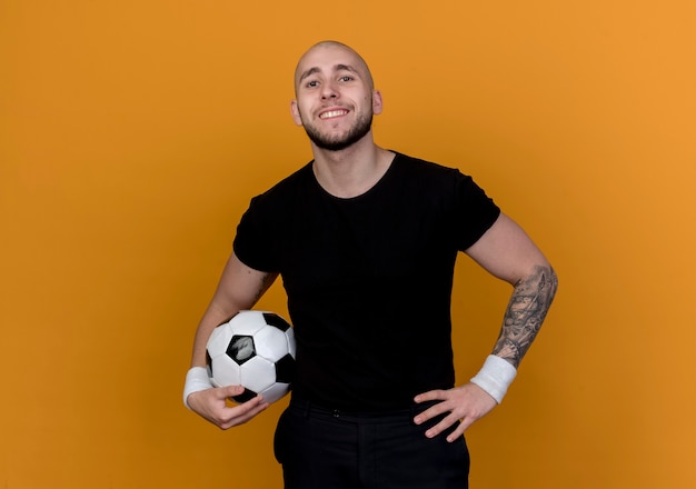 Lächelnder junger sportlicher Mann, der Armband hält Ball hält und Hand auf Hüfte lokalisiert auf orange Wand setzt