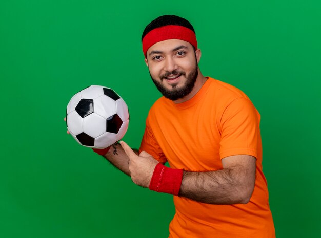 Lächelnder junger sportlicher Mann, der Stirnband und Armband hält und Punkte auf Kugel lokalisiert auf grünem Hintergrund trägt