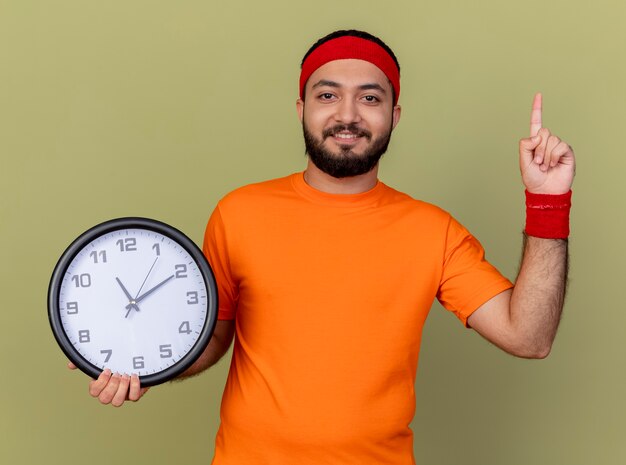 Lächelnder junger sportlicher Mann, der Stirnband und Armband hält Wanduhr und zeigt oben auf lokalisiert auf olivgrünem Hintergrund