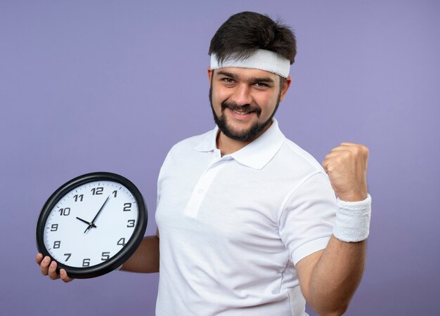 Lächelnder junger sportlicher Mann, der Stirnband und Armband hält Wanduhr zeigt ja Geste