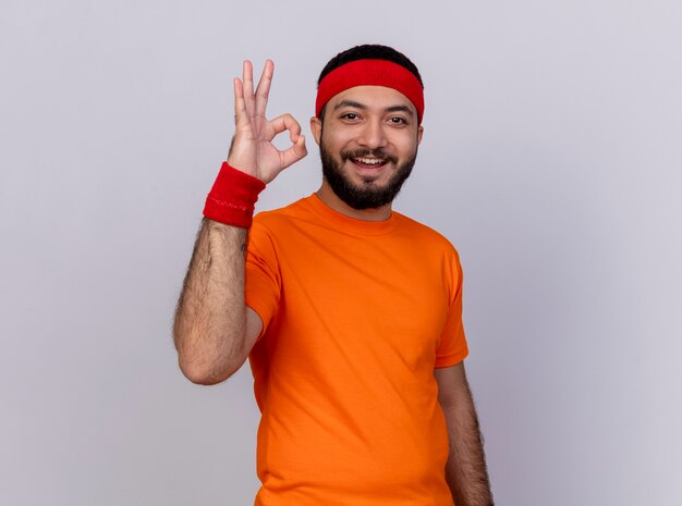 Lächelnder junger sportlicher Mann, der Stirnband und Armband trägt, zeigt okay Geste lokalisiert auf Weiß