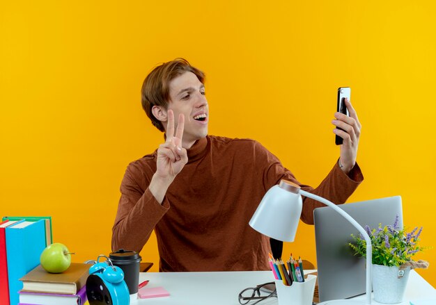 lächelnder junger Studentenjunge, der mit Schulwerkzeugen am Schreibtisch sitzt, macht ein Selfie und zeigt Friedensgeste