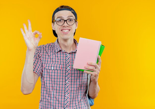 Lächelnder junger Studentenjunge, der Rückentasche und Brille und Kappe hält, Notizbuch hält und okey Geste auf Weiß zeigt