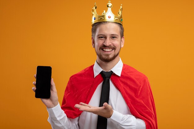 Lächelnder junger Superheldenmann, der Krawatte und Krone hält und Punkte mit Hand am Telefon lokalisiert auf orange Hintergrund trägt